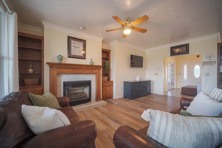 Living area featuring wood-finish flooring, a fireplace with a wood mantle and tile surround, built-in wooden shelving, crown molding, and a ceiling fan