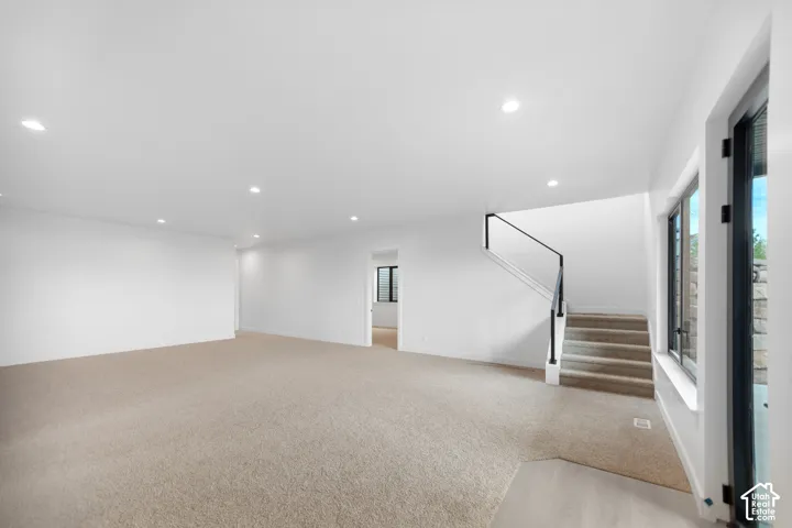 Spacious room with recessed lighting, light-toned carpeting, staircase with black railing, and large window