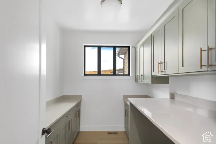 White walls with a black-framed window, light wood-finish flooring, and light gray cabinetry with gold-tone hardware