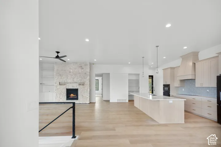 Open-concept living space with light wood-finish flooring, a stone fireplace, built-in shelving, a kitchen island with pendant lighting, and light cabinetry with a tile backsplash