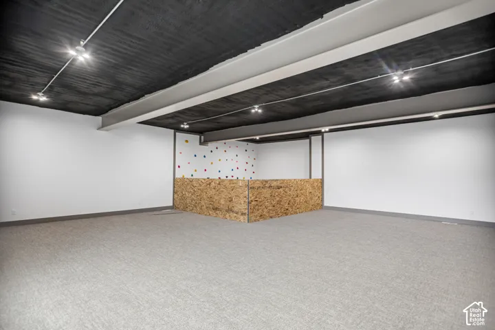 Spacious room featuring textured gray carpeting, white walls, exposed ceiling beams, track lighting, and an oriented strand board half-wall