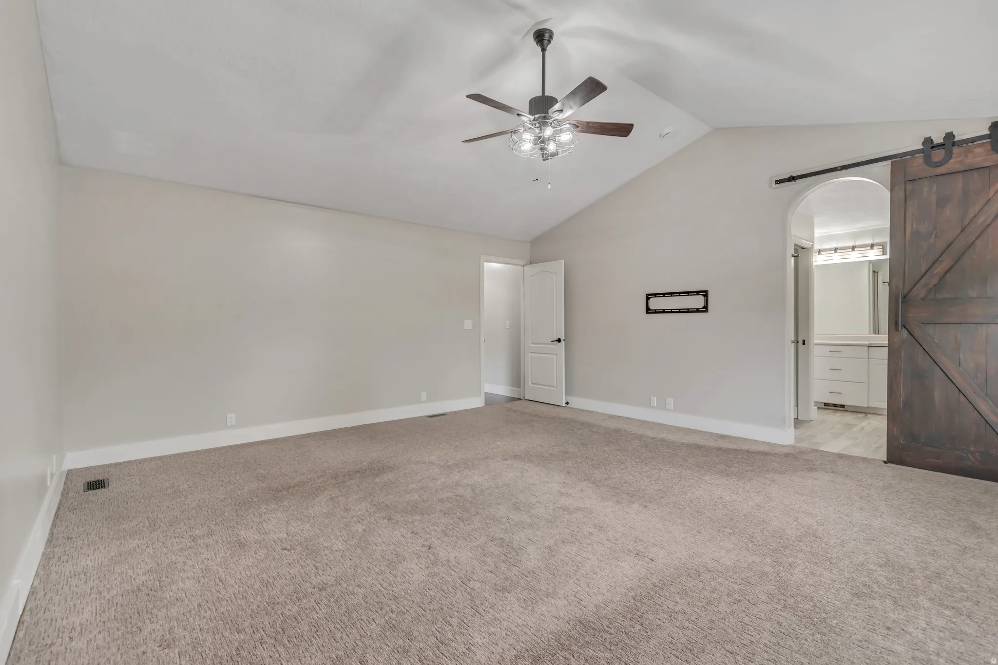 Unfurnished bedroom with a barn door, lofted ceiling, light colored carpet, ceiling fan, and ensuite bathroom