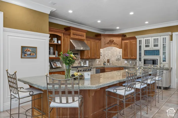 Kitchen featuring brown cabinetry, tasteful backsplash, a breakfast bar area, wall chimney range hood, and light tile patterned floors