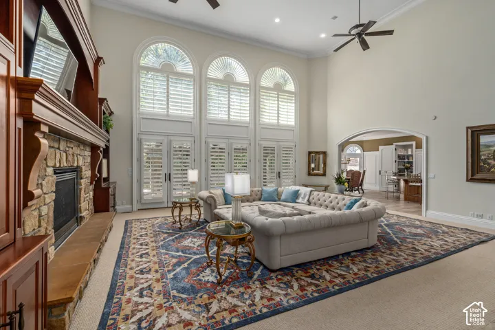 Living area featuring a ceiling fan, carpet, a towering ceiling, ornamental molding, and a stone fireplace