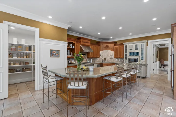 Kitchen featuring brown cabinets, recessed lighting, light tile patterned flooring, light stone counters, and crown molding