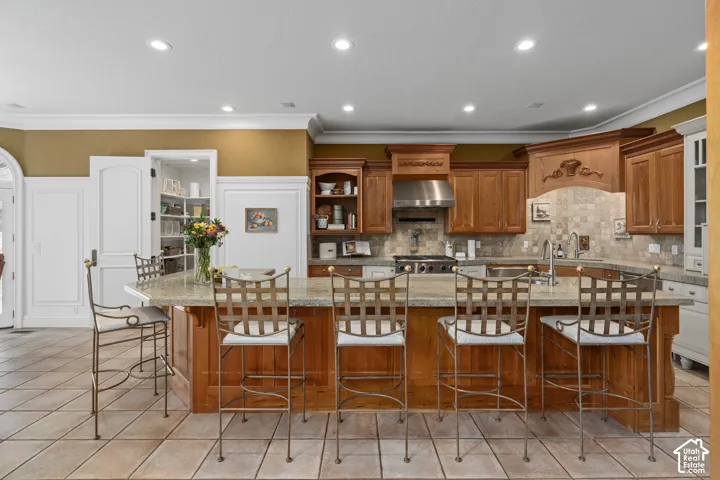 Kitchen with brown cabinetry, a kitchen bar, light stone counters, crown molding, and light tile patterned floors