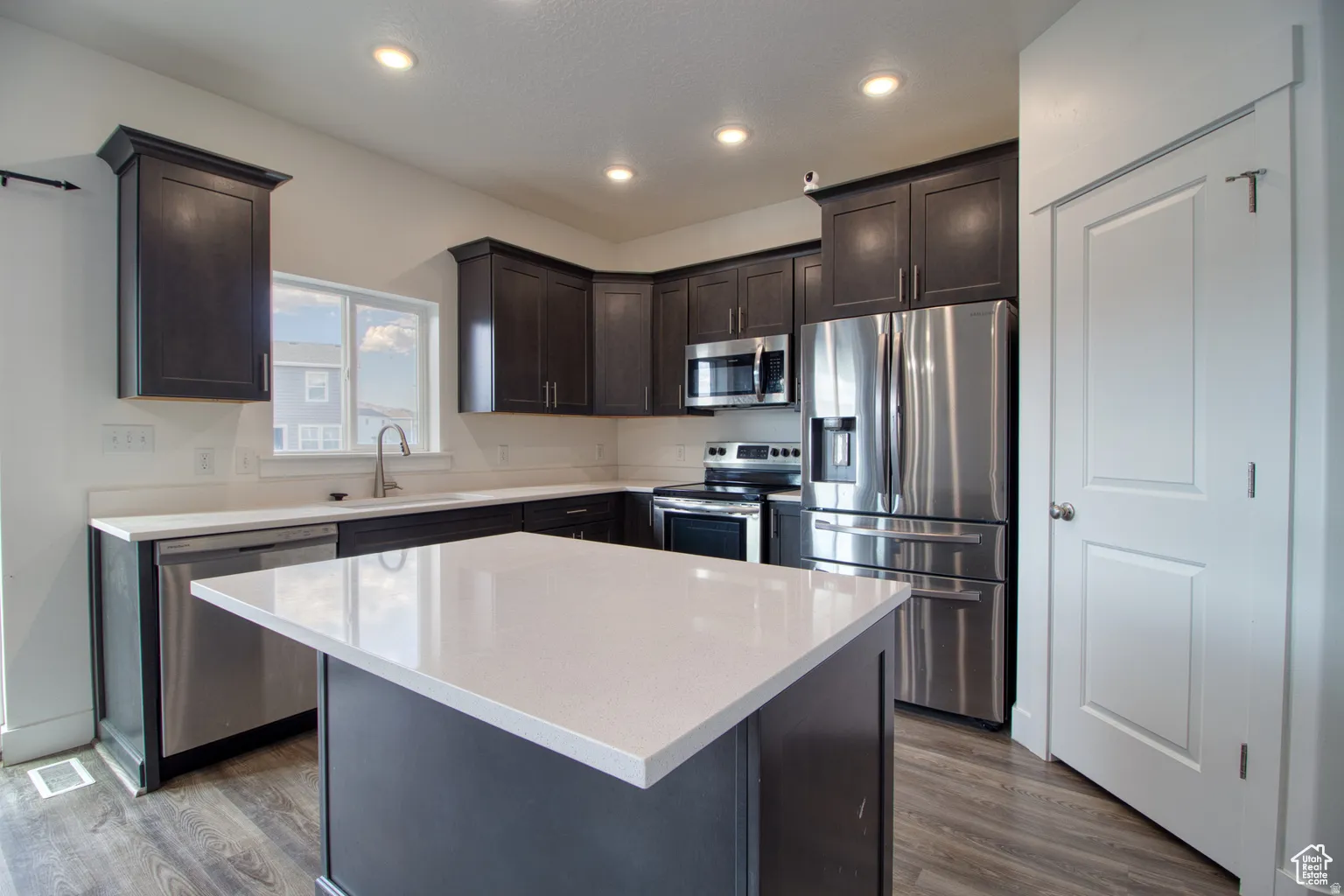 Kitchen with appliances with stainless steel finishes, dark brown cabinetry, dark wood-style flooring, light stone counters, and recessed lighting