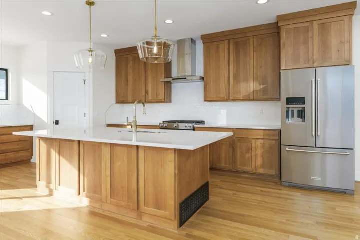 Kitchen with brown cabinets, stainless steel appliances, wall chimney range hood, decorative backsplash, and light wood finished floors