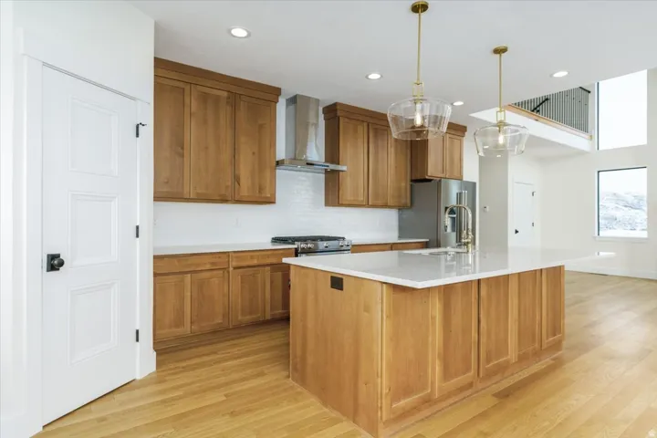 Kitchen with brown cabinetry, wall chimney range hood, backsplash, a center island with sink, and recessed lighting