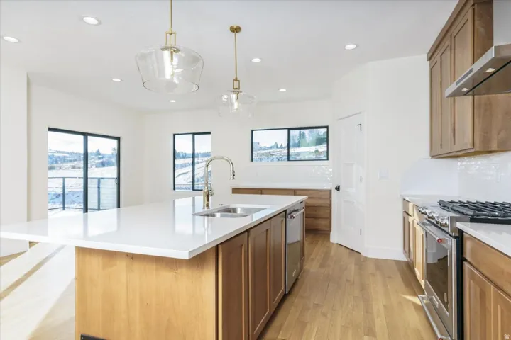 Kitchen with stainless steel gas range oven, decorative light fixtures, wall chimney exhaust hood, light wood-style floors, and light stone counters