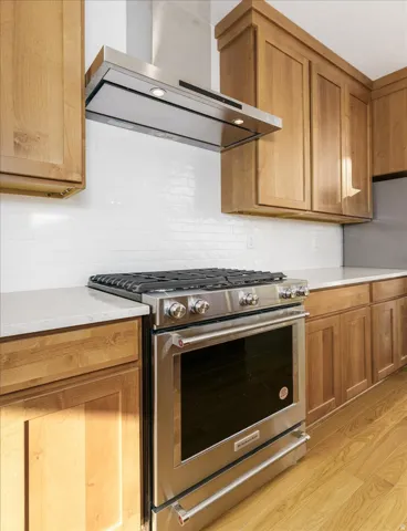 Kitchen with stainless steel range with gas cooktop, exhaust hood, light wood finished floors, light stone counters, and brown cabinets