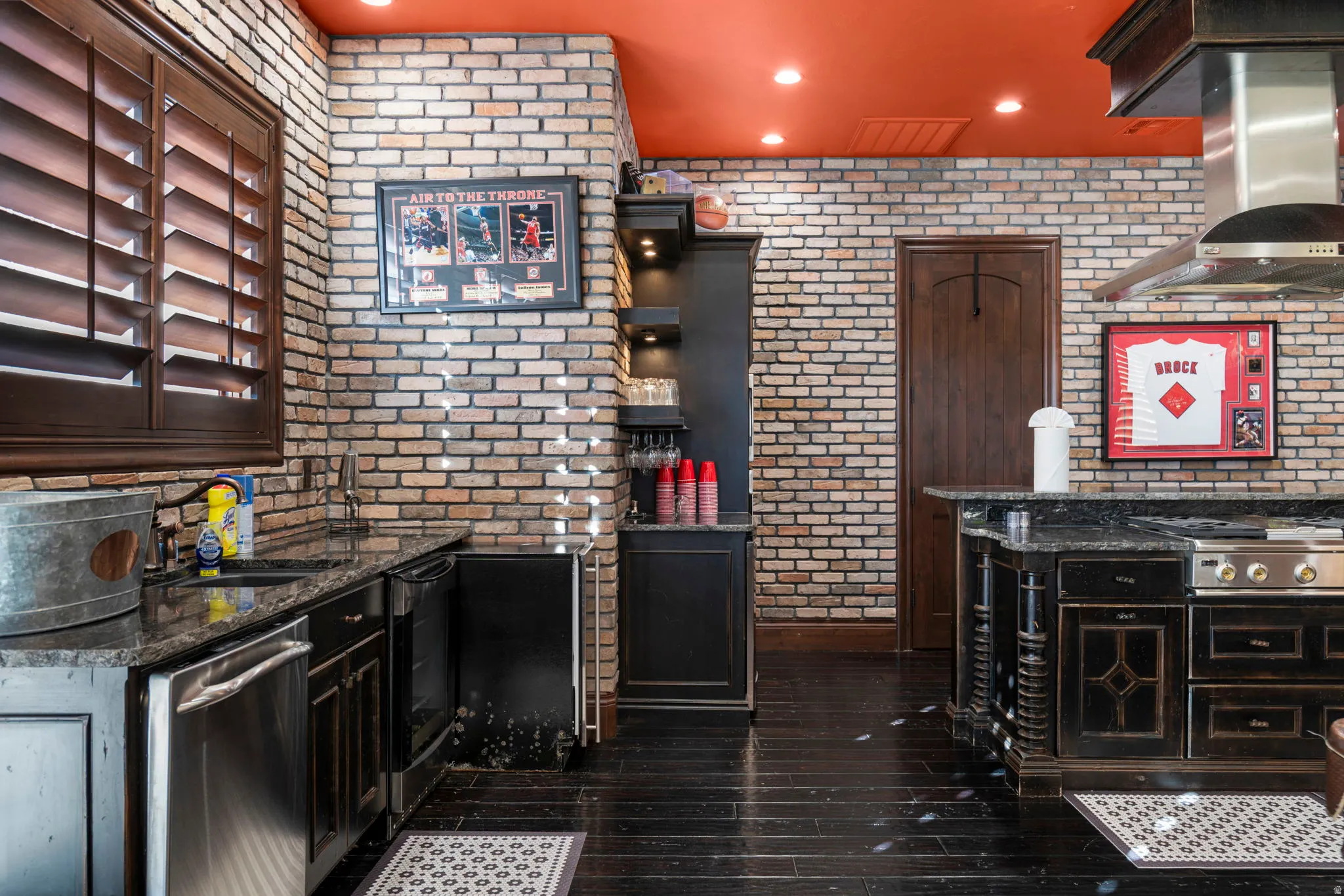 Kitchen with dark stone countertops, wall chimney range hood, brick wall, dishwasher, and dark wood-type flooring