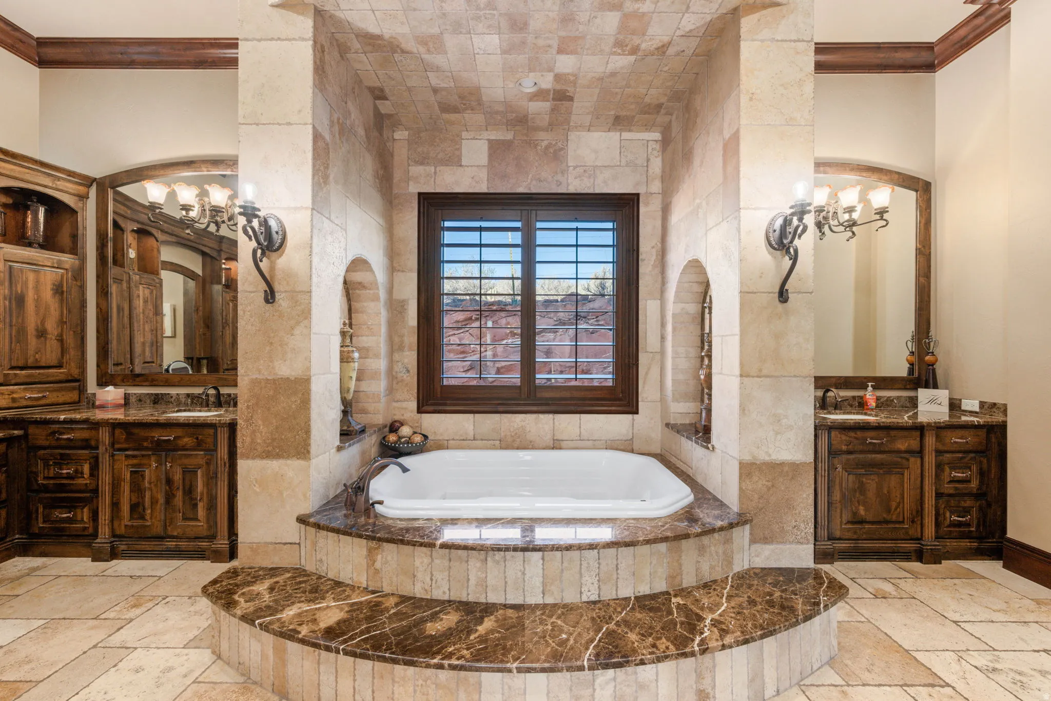 Full bathroom featuring stone tile flooring, two vanities, a garden tub, crown molding, and a chandelier