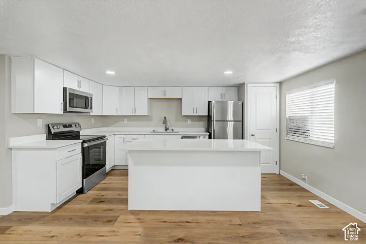 Kitchen featuring white cabinetry, stainless steel appliances, wood-finish flooring, a central island, and a window with blinds