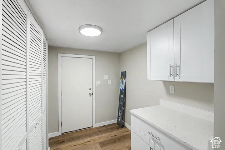 White louvered closet doors, wood-finish flooring, white flat-panel cabinetry, light speckled countertop, and a flush-mount ceiling light fixture