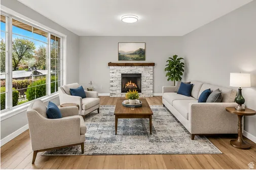 Spacious living area featuring large picture windows, wood-finish flooring, a stone-clad fireplace with a wood mantle, and a flush-mount ceiling light