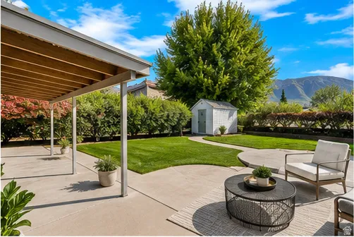 Expansive concrete patio with covered area, manicured lawn, storage shed, and mature trees