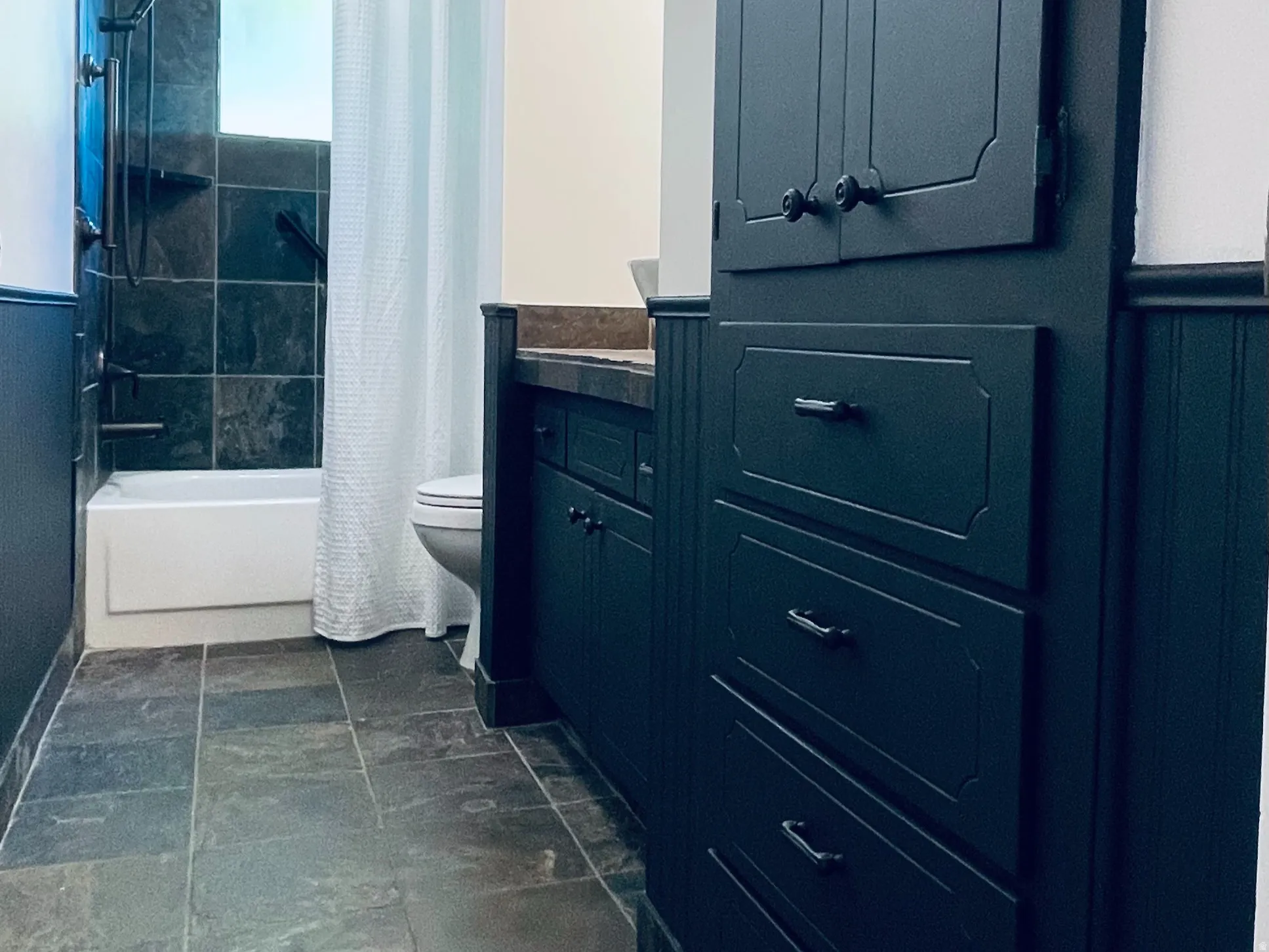 Bathroom featuring dark slate floor tiles, a built-in dark wood cabinetry unit with varied storage, dark wood wainscoting, and a white tub with dark tiled surround