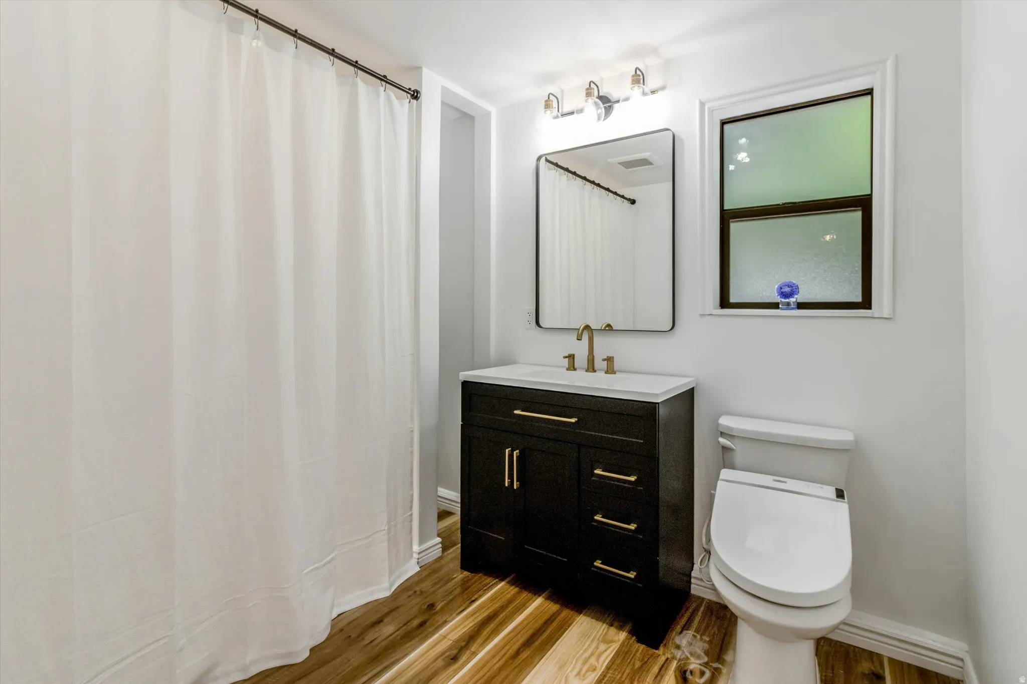 Bathroom featuring a dark wood-finish vanity with a white countertop and gold-tone fixtures, a rectangular vanity mirror, and wood-finish flooring