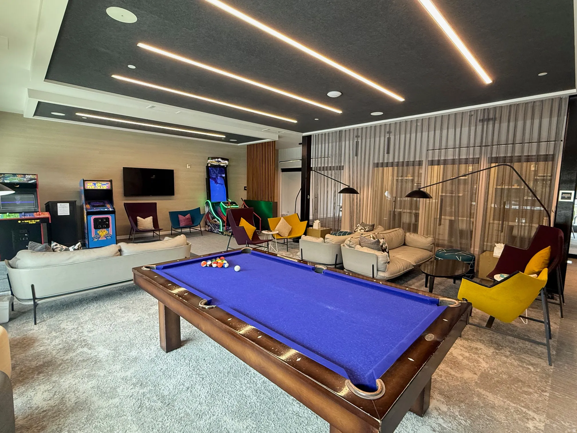 Spacious recreational area featuring a billiard table, integrated linear ceiling lighting, a wall-mounted display, and large windows with sheer privacy treatments