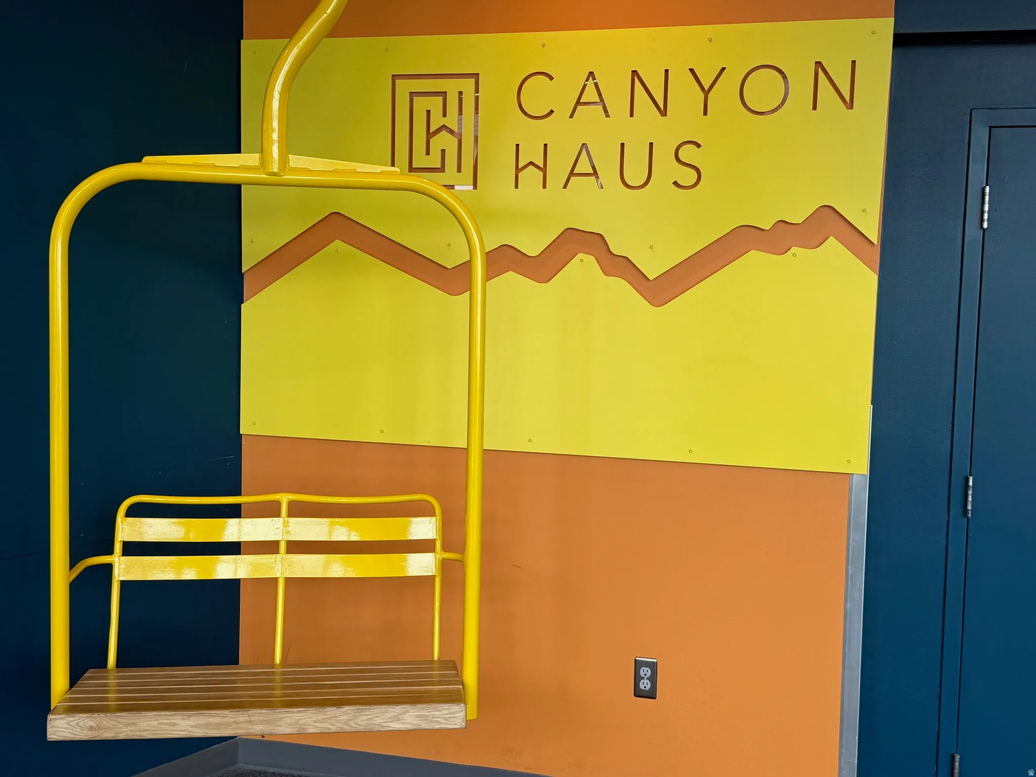 Ski lift chair with a wood-finish seat, set against a two-tone wall featuring orange and blue sections