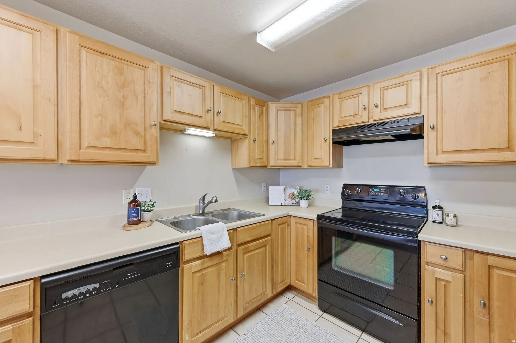 Kitchen featuring light wood cabinetry, light-toned countertops, a double basin stainless steel sink, black electric range, and black dishwasher