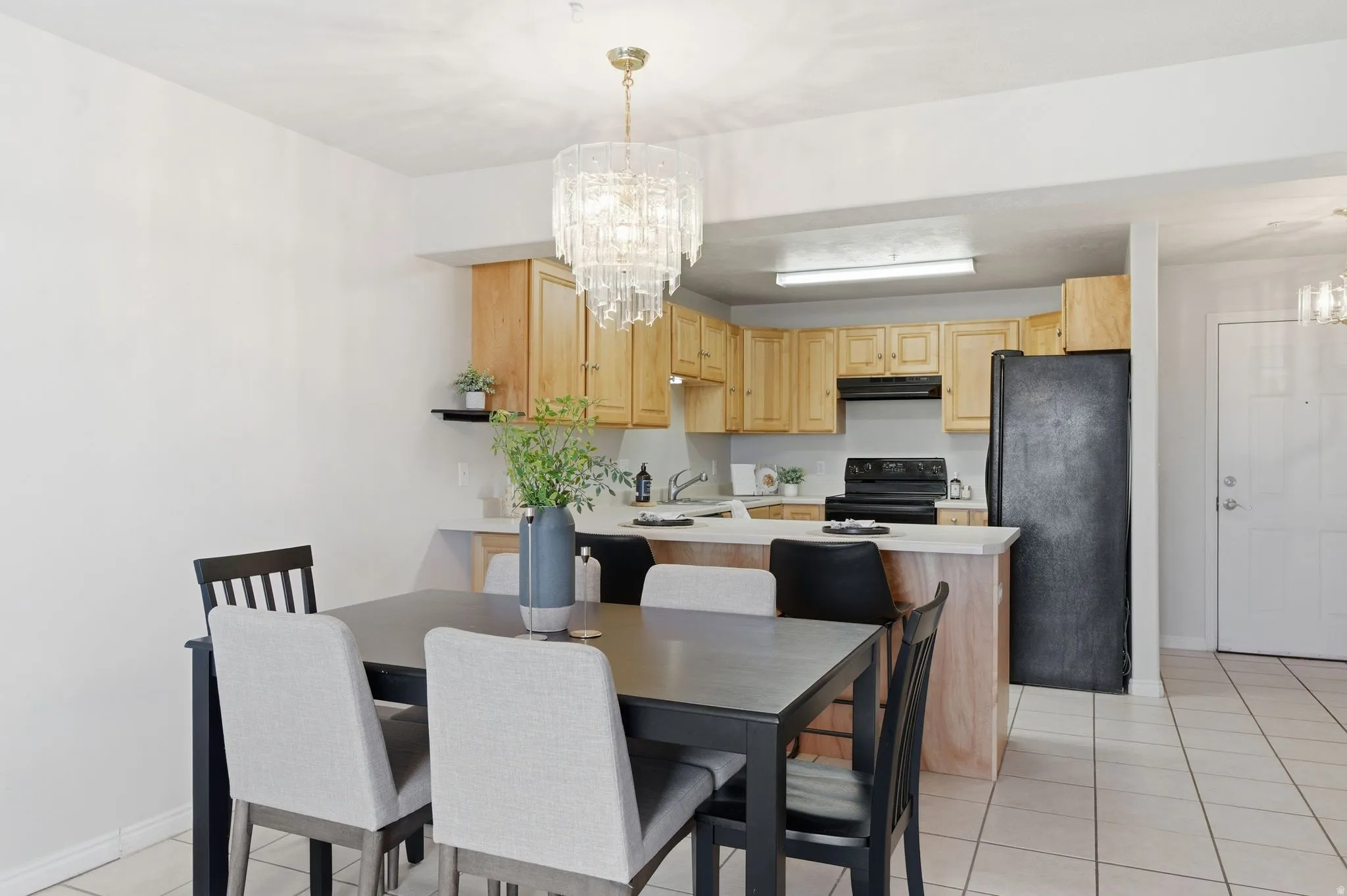 Dining area with a sophisticated crystal chandelier, tile flooring, and a kitchen featuring light wood cabinetry, a breakfast bar, and black appliances