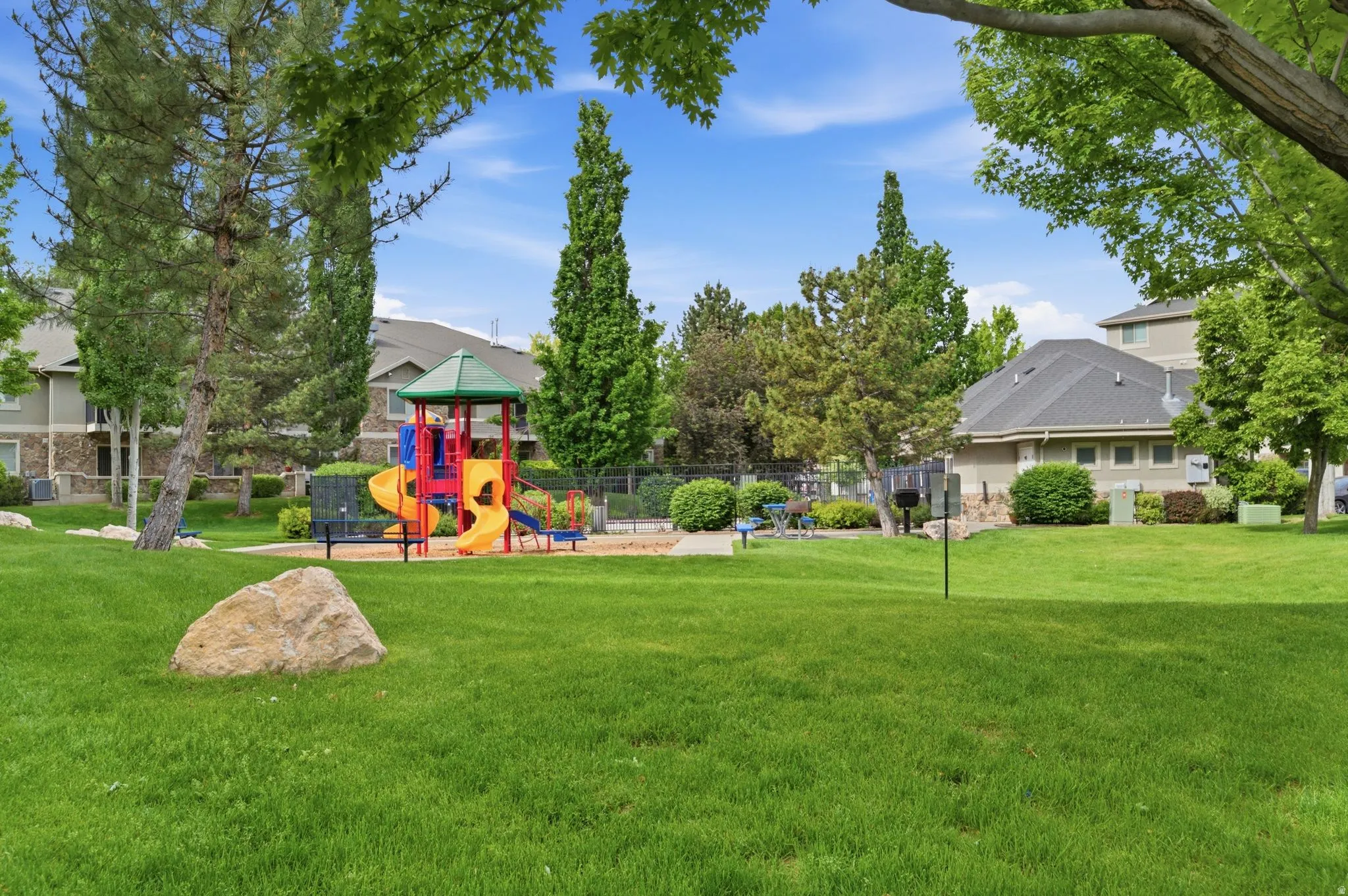 Community green space featuring a vibrant playground structure with multiple slides and climbing elements, surrounded by mature trees