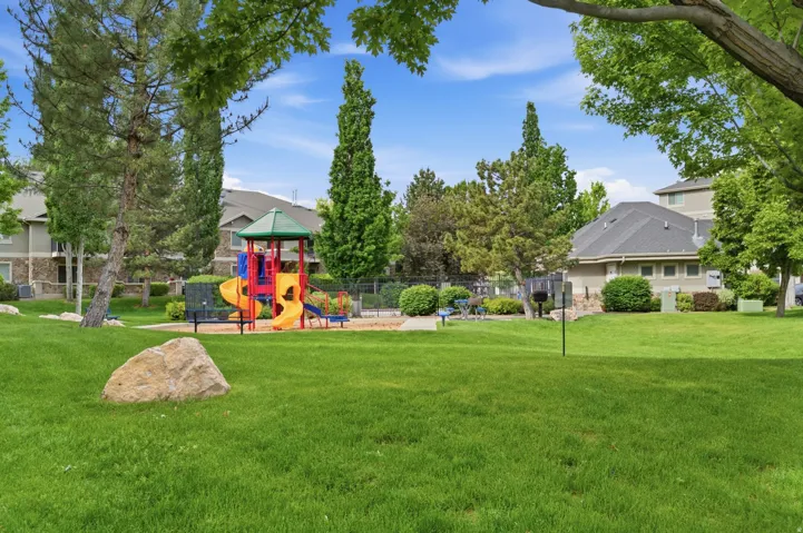 Community green space featuring a vibrant playground structure with multiple slides and climbing elements, surrounded by mature trees