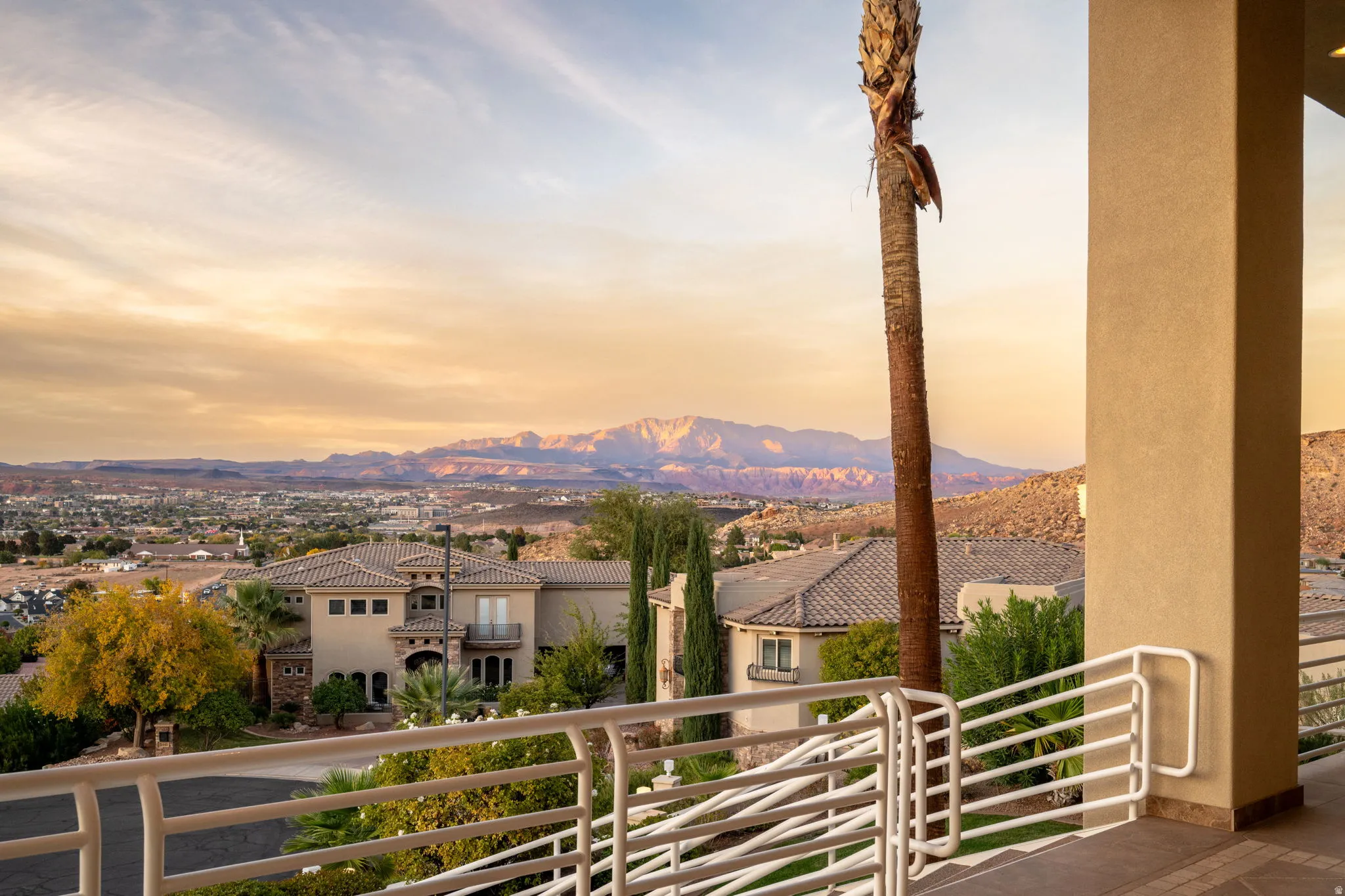 Expansive outdoor balcony with white horizontal railings and stucco columns, overlooking a residential area with distant mountain ranges