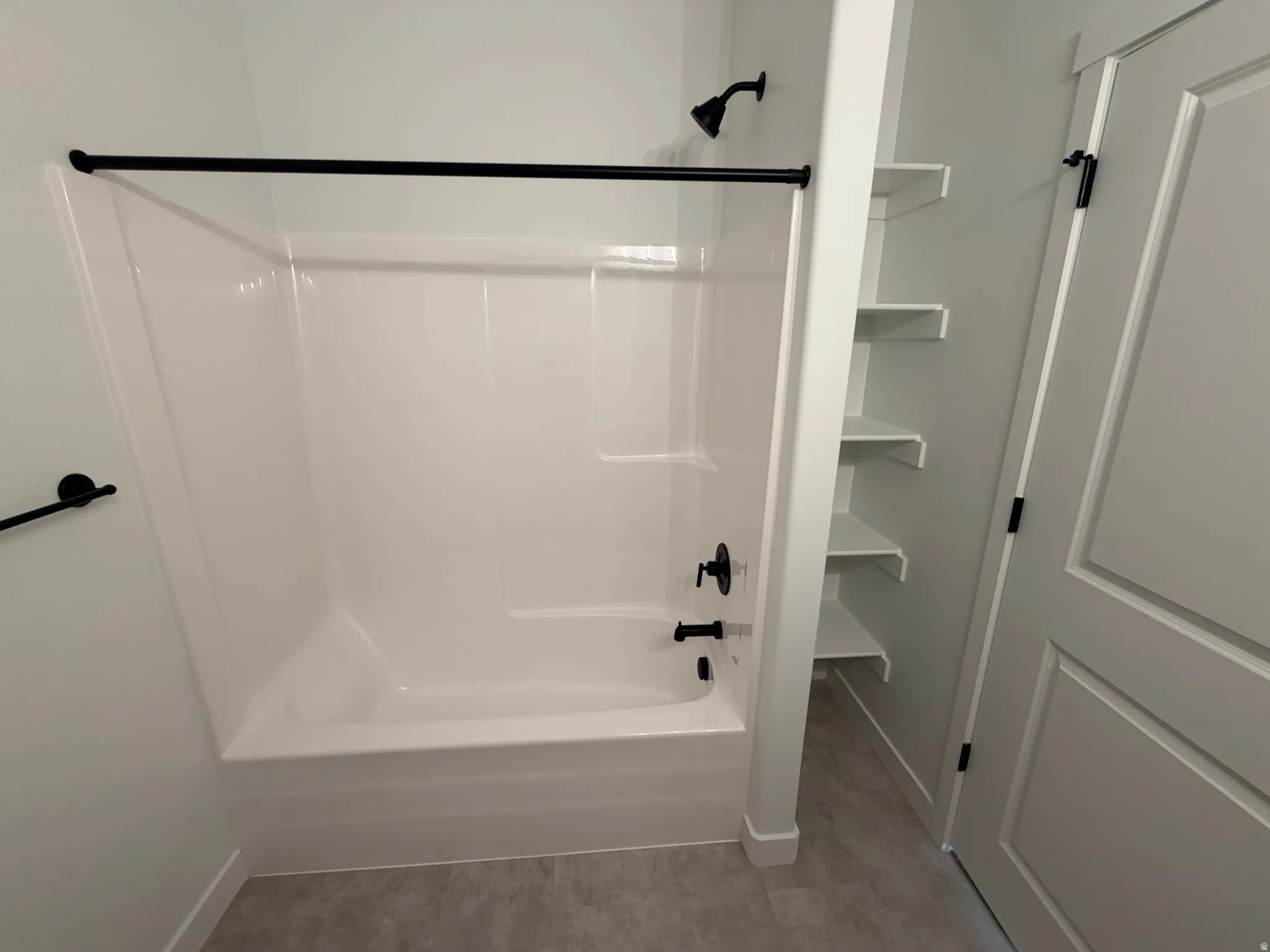Bathtub/shower combination featuring a matte black shower head and faucet, alongside built-in open shelving and a paneled door with black hardware