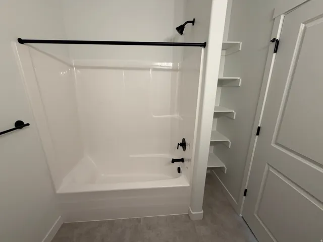 Bathtub/shower combination featuring a matte black shower head and faucet, alongside built-in open shelving and a paneled door with black hardware