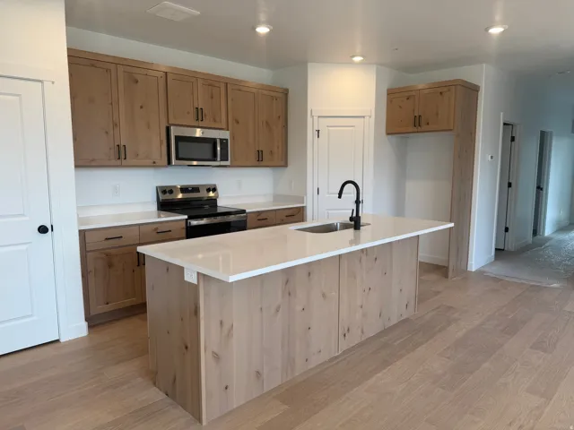 Kitchen featuring wood-finish cabinetry, stainless steel appliances, a center island with sink and matte black faucet, light wood-finish flooring, and recessed lighting