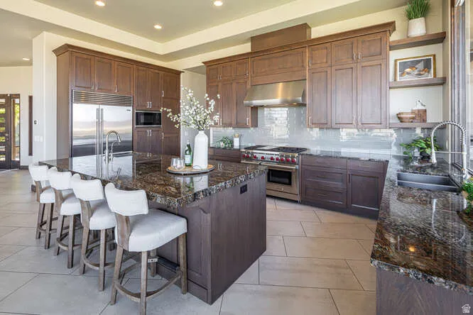 Kitchen featuring decorative backsplash, open shelves, built in appliances, an island with sink, and range hood