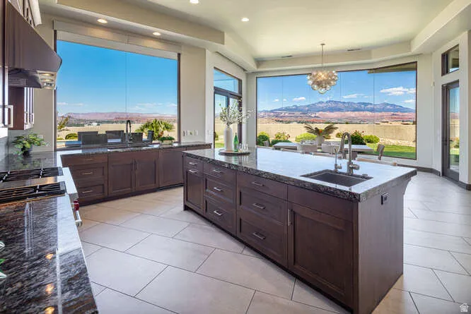 Kitchen with dark stone countertops, hanging light fixtures, an island with sink, under cabinet range hood, and dark brown cabinets