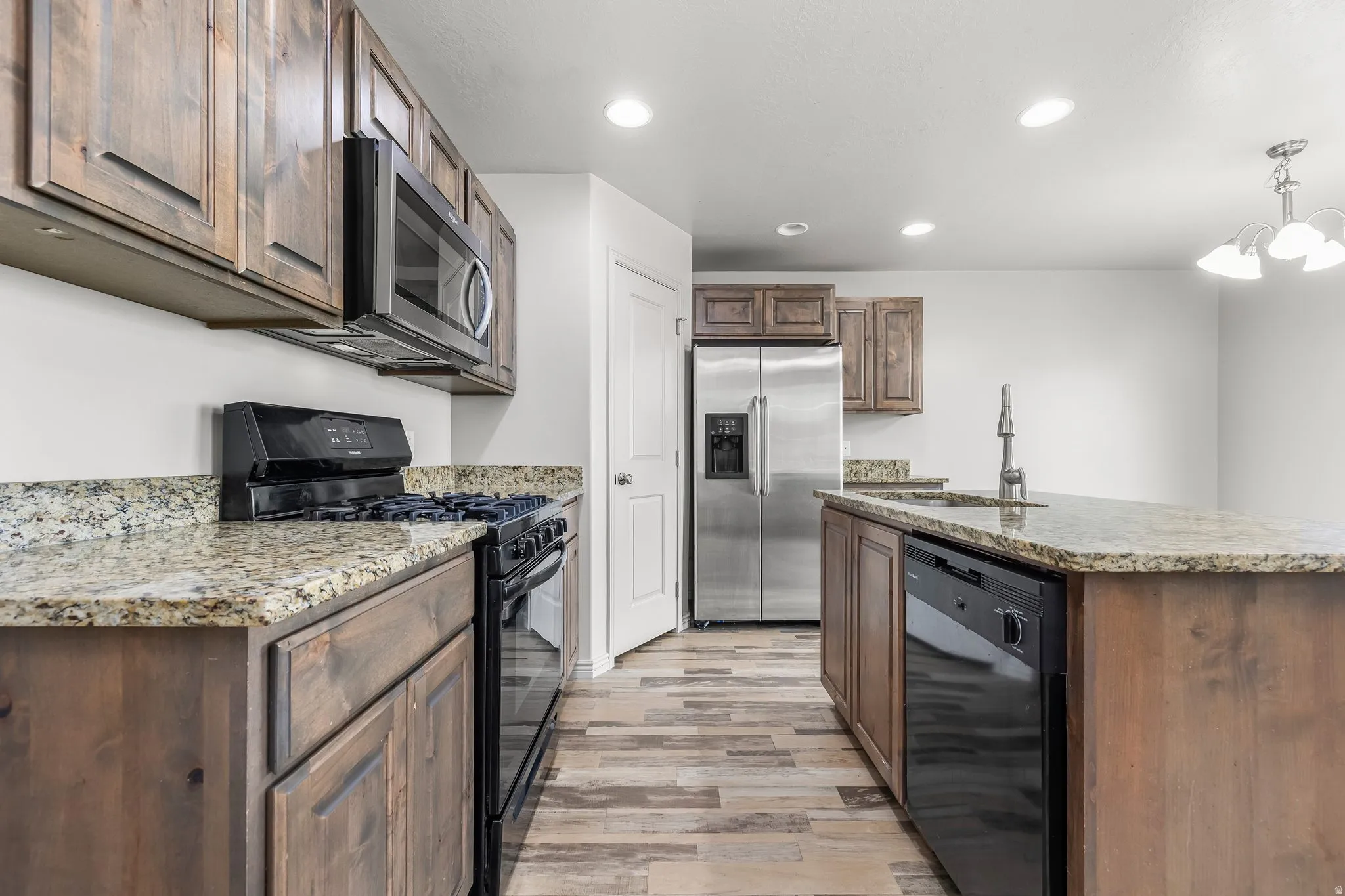 Kitchen featuring black appliances, recessed lighting, light wood finished floors, light stone countertops, and an island with sink