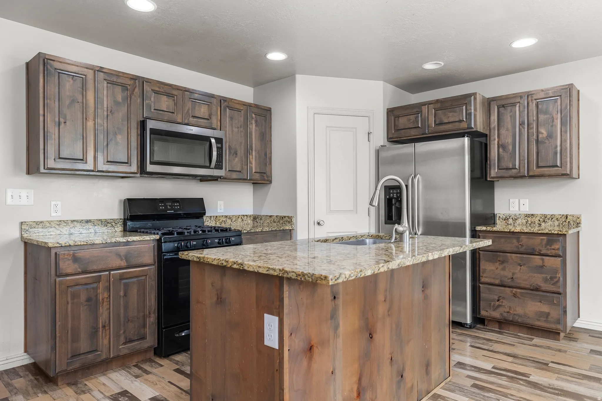 Kitchen featuring appliances with stainless steel finishes, dark brown cabinetry, light stone counters, recessed lighting, and an island with sink