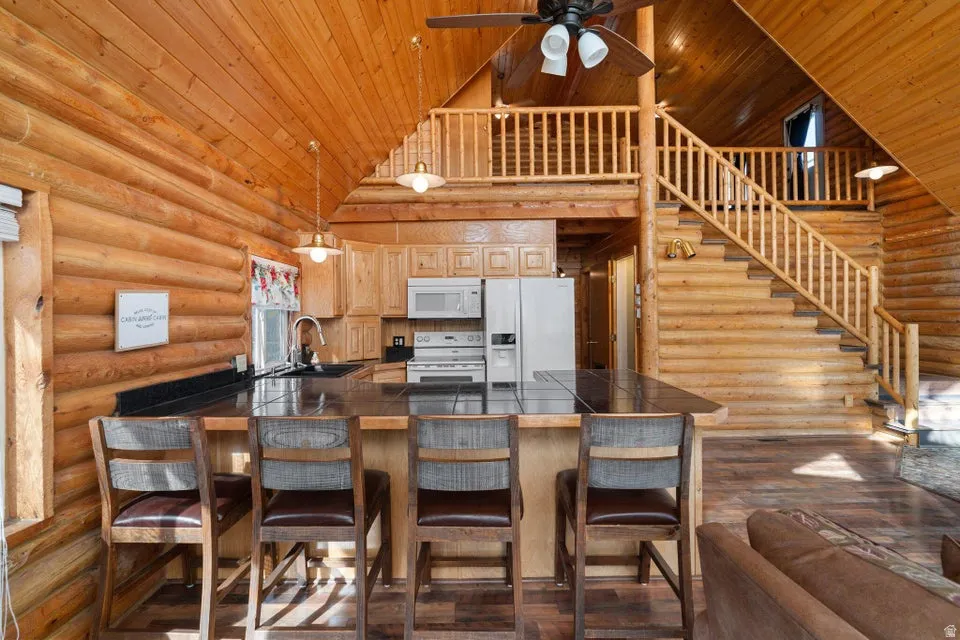 Kitchen featuring log walls, high vaulted ceiling, a peninsula, white appliances, and hanging light fixtures