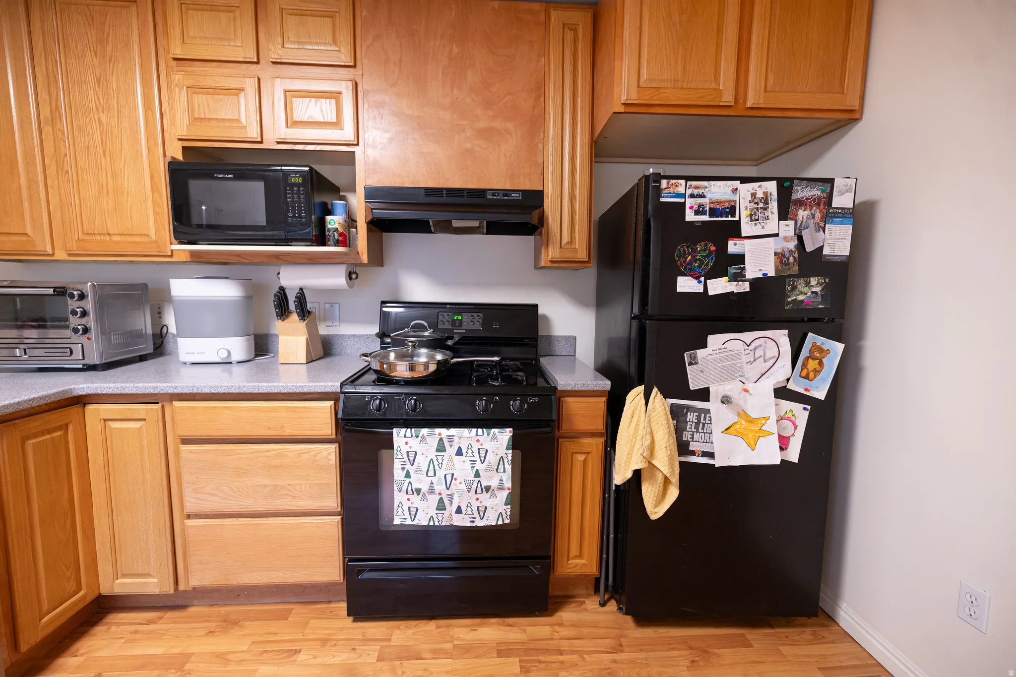 Kitchen featuring black appliances, light countertops, light wood-style flooring, under cabinet range hood, and brown cabinetry