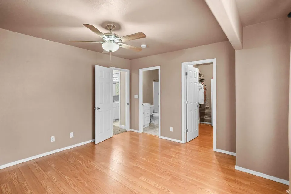 Unfurnished bedroom with a walk in closet, light wood finished floors, ceiling fan, connected bathroom, and beamed ceiling