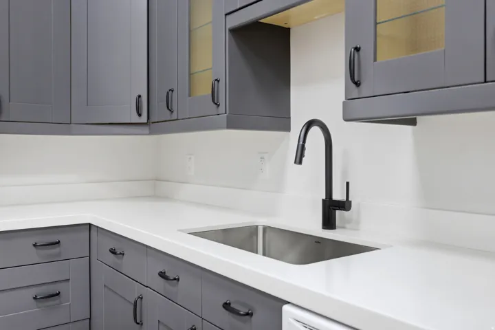 Contemporary kitchen featuring gray shaker-style cabinetry, white countertops, a stainless steel undermount sink, a matte black pull-down faucet, and upper glass-front cabinets