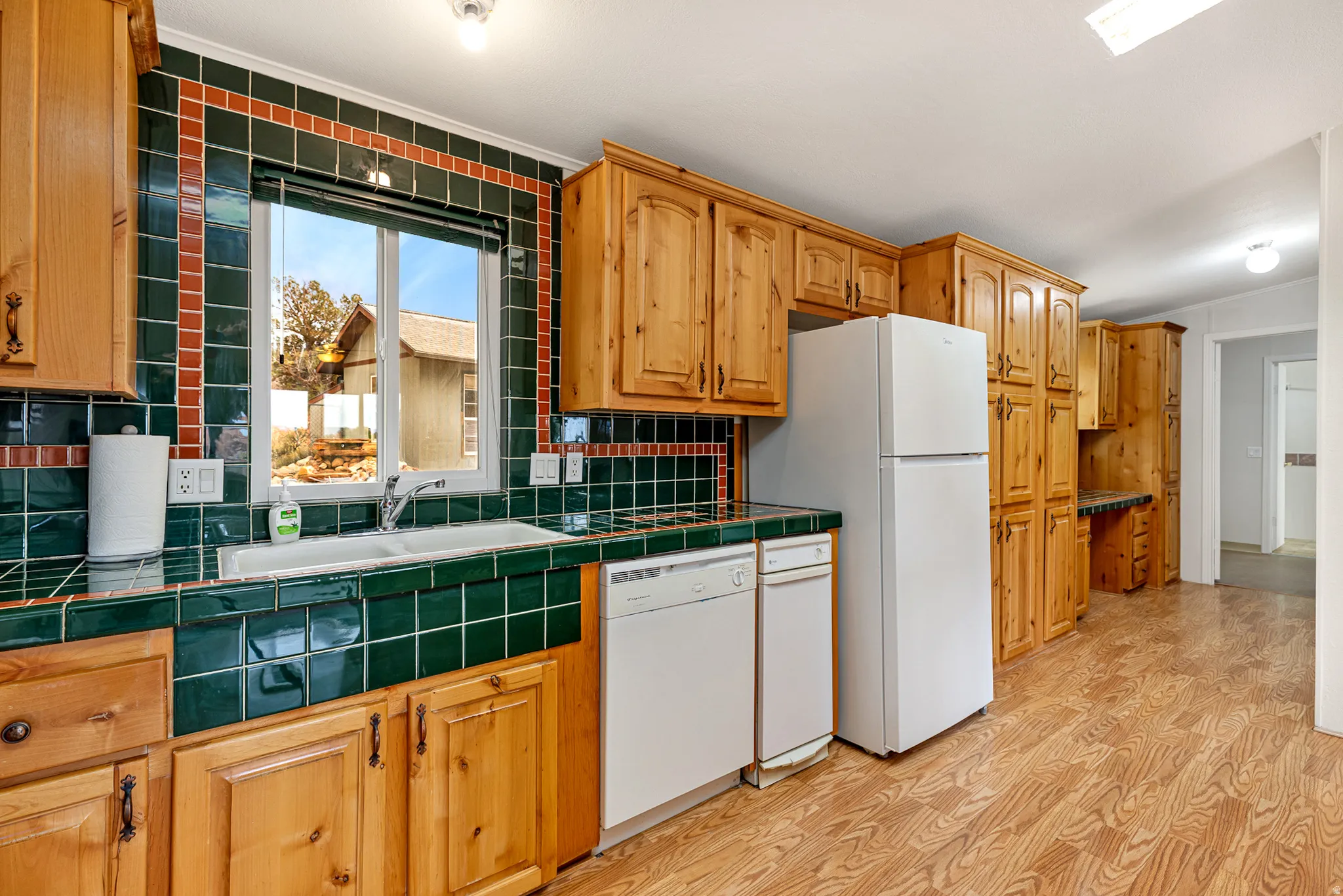 Kitchen with tile counters, tasteful backsplash, white appliances, light wood finished floors, and brown cabinetry