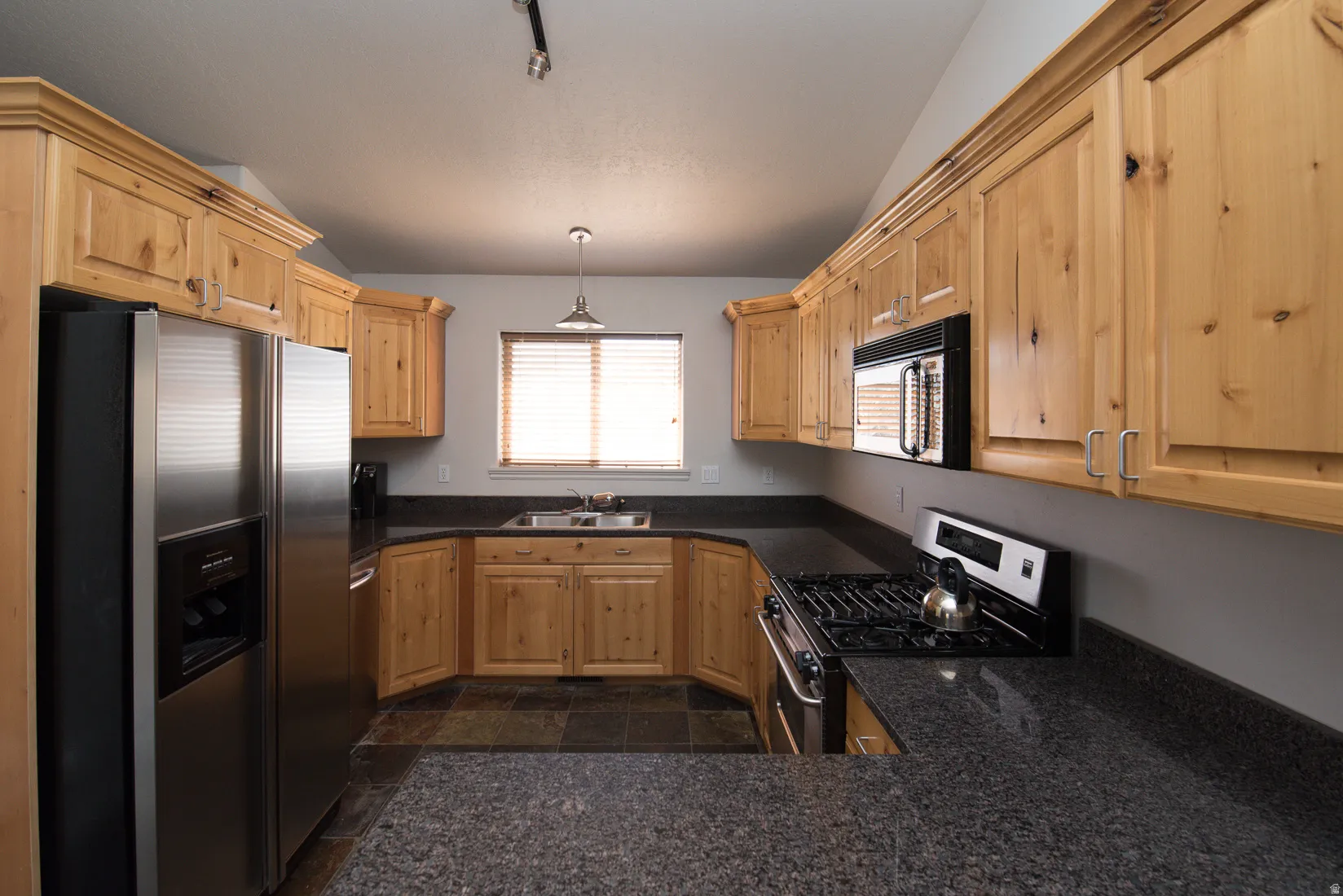 Kitchen featuring appliances with stainless steel finishes, hanging light fixtures, light brown cabinetry, lofted ceiling, and dark stone counters