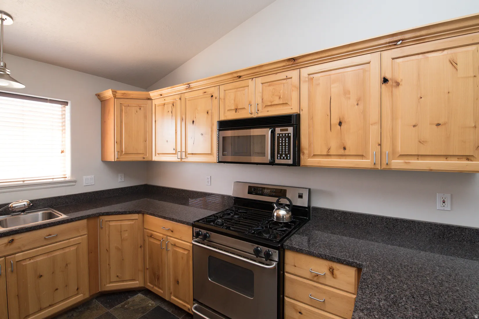 Kitchen featuring stainless steel appliances, lofted ceiling, decorative light fixtures, light brown cabinetry, and dark stone countertops