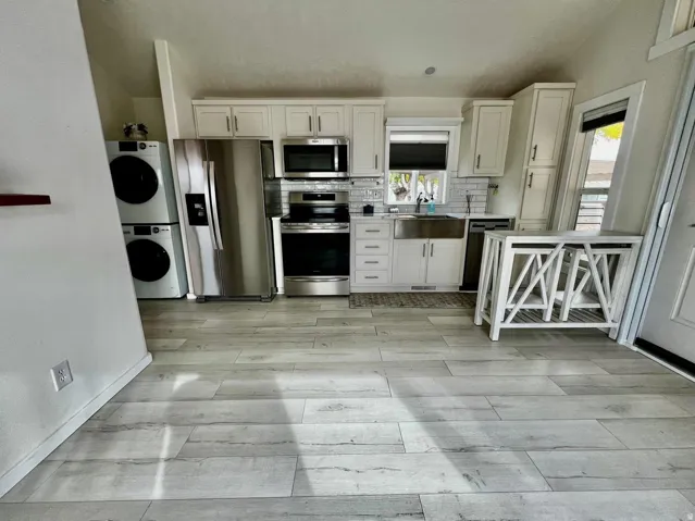 Kitchen with appliances with stainless steel finishes, backsplash, white cabinets, light wood-style floors, and estacked washer and dryer