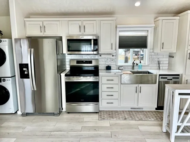 Kitchen featuring stainless steel appliances, white cabinetry, light stone countertops, stacked washer / drying machine, and recessed lighting