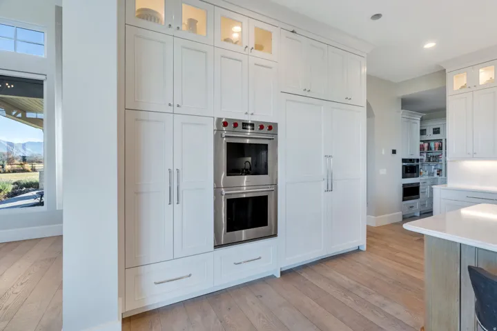 Kitchen with glass insert cabinets, stainless steel double oven, white cabinetry, light wood finished floors, and light stone countertops