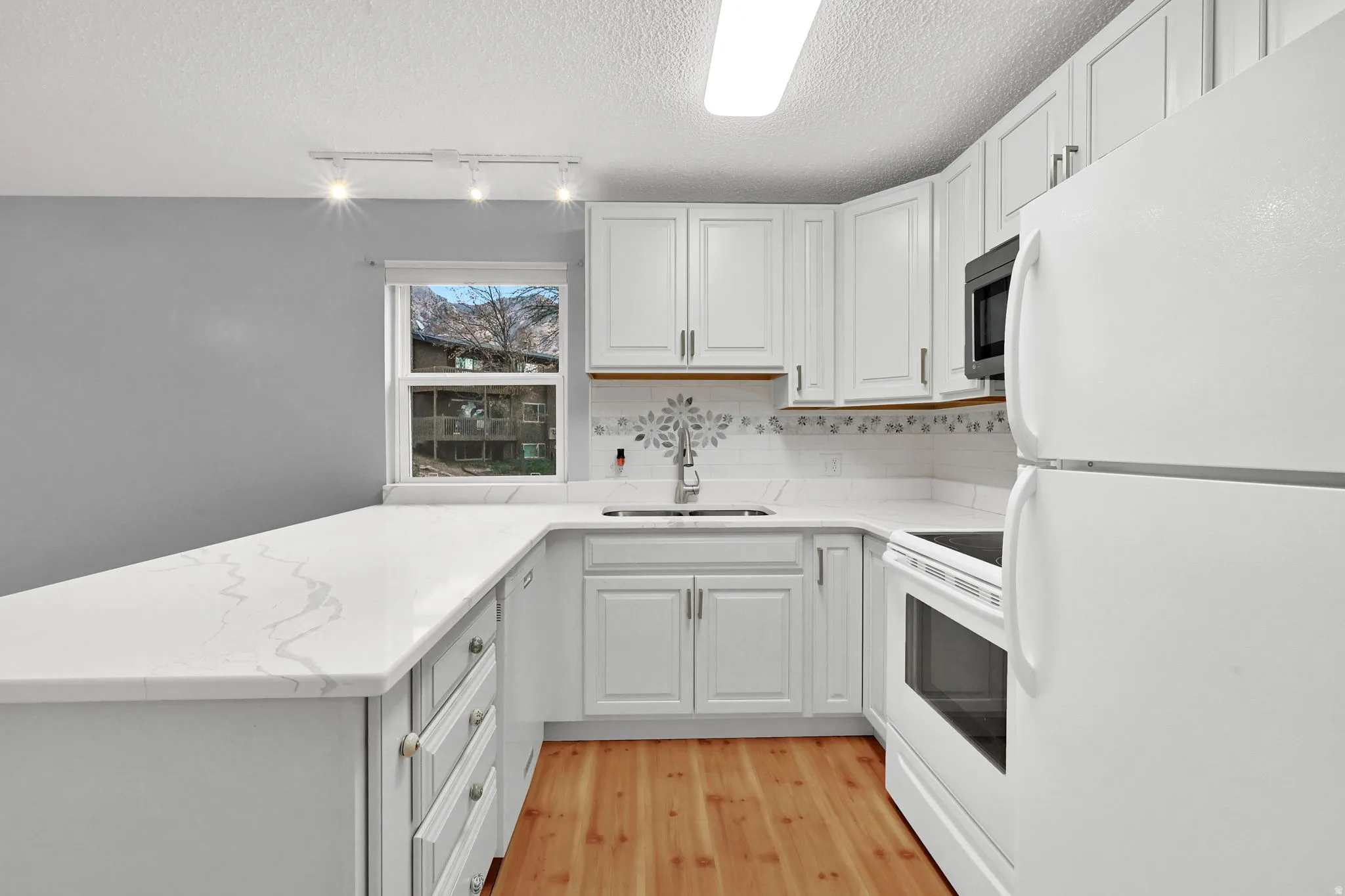 Kitchen with white appliances, a peninsula, light wood-type flooring, rail lighting, and white cabinets