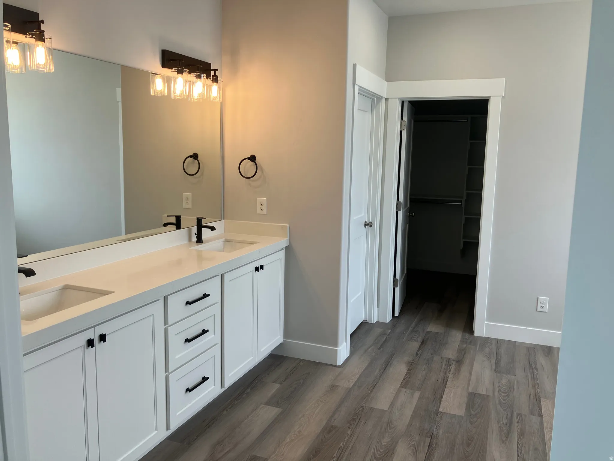 Dual-vanity primary bathroom featuring white cabinetry with multiple drawers, quartz countertops, vessel sinks, matte black hardware and fixtures, framed mirrors with integrated lighting, and wood-finish plank flooring throughout