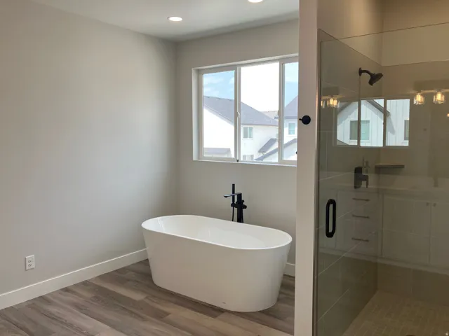 Spa-inspired primary bathroom featuring a freestanding oval soaking tub with matte black deck-mounted faucet, frameless glass shower enclosure with tile surround, wood-finish flooring, recessed ceiling lighting, and natural light from a large window overl